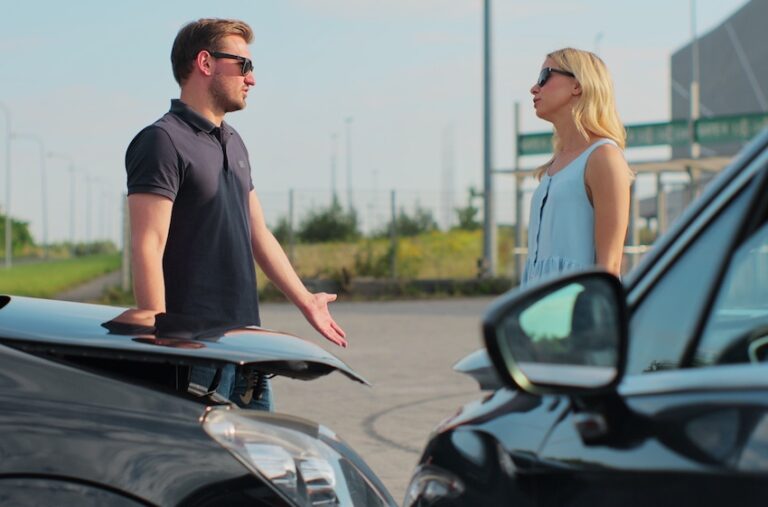 Two drivers in sunglasses are discussing and gesturing near their damaged vehicles on the roadside after a shared fault accident collision in Georgia.