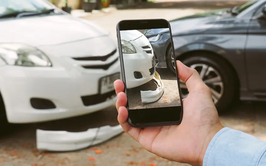 A person is holding a smartphone and is photographing crushed front-end damage between the white and black cars and is documenting the shared fault accident scene in Georgia.