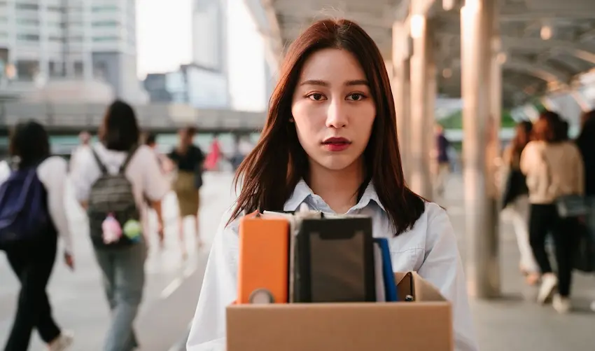 A young Asian woman with a depressed expression carries a box of her belongings out of her workplace representing an employee who has been wrongfully terminated after filing for workers' compensation leave in Georgia.