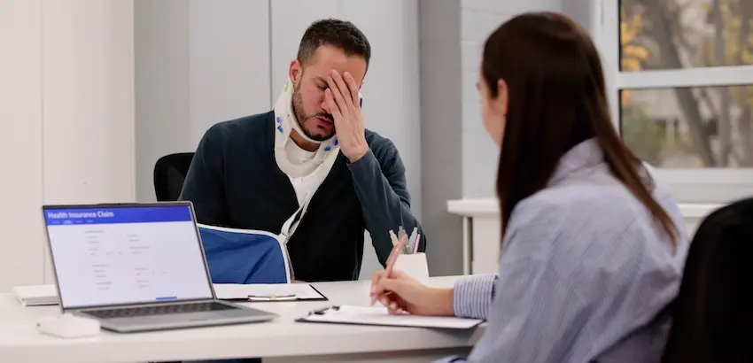A distressed man wearing a neck brace and arm sling holds his hand to his face while sitting across from a woman reviewing documents beside a laptop representing a workplace injury victim meeting with a lawyer to discuss his want to switch his workers' comp physician at the Law Offices of Humberto Izquierdo Jr. PC in Georgia.