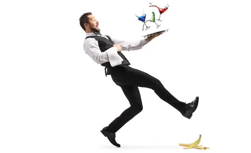 A waiter in uniform loses his balance and slips on a banana peel while carrying a tray of cocktails illustrating the type of workplace accident that would require an injured employee to be evaluated by a workers' comp physician in Georgia.