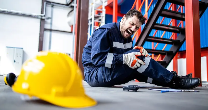A male warehouse worker in a safety uniform sits on the floor grimacing in pain while clutching his knee beside a fallen yellow hard hat depicting a workplace injury that would qualify an employee for workers' compensation leave in Georgia.