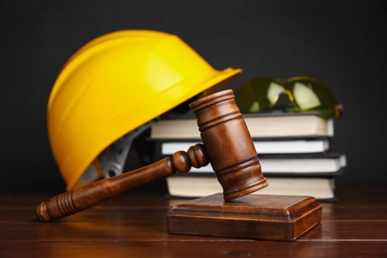A wooden judge's gavel resting on its sound block in front of a yellow construction hard hat safety glasses and a stack of books symbolizing the legal process involved in filing a workers' compensation leave claim after a workplace injury in Georgia.