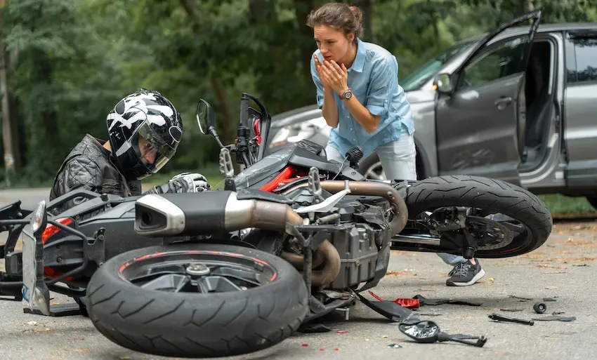 A motorcycle rider in a helmet and riding gear lies on the pavement next to a downed bike with debris scattered across the road while a visibly shaken woman in a light blue shirt crouches over him with her hands raised to her mouth representing the devastating aftermath of an uninsured motorist striking a motorcyclist in Georgia.