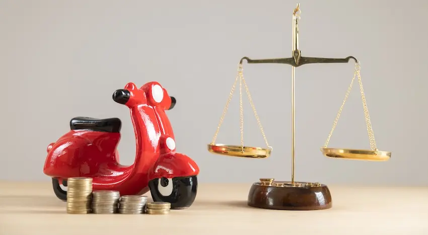 A red ceramic scooter figurine sitting beside two stacks of coins and a gold balance scale on a wooden surface symbolizing the financial and legal weight of pursuing compensation after being hit by an uninsured motorist with help from the Law Offices of Humberto Izquierdo Jr. PC.