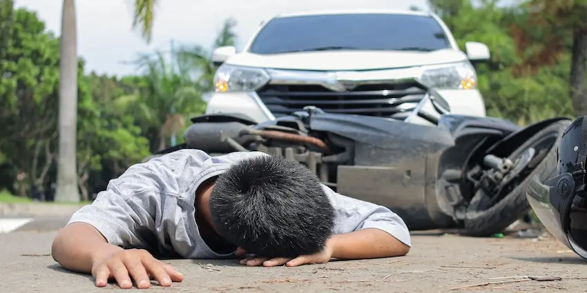 A motorcycle rider in a gray shirt lies face-down and motionless on a road with his helmet beside him and his wrecked motorcycle visible in the background as a white SUV with front-end damage sits stopped at the scene of a collision caused by an uninsured motorist in Georgia.