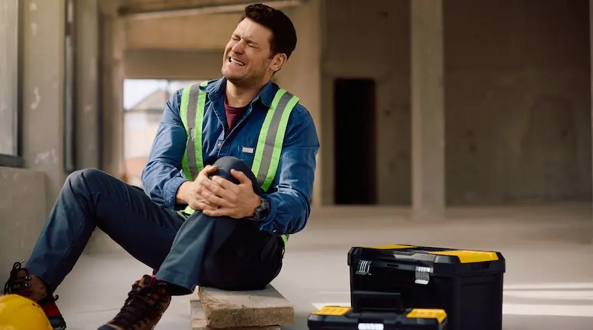 A construction worker in a safety vest sits on the floor of a job site, grimacing and clutching his injured knee beside his toolbox depicting the type of on-the-job injury that leads to a workers' comp claim in Georgia.