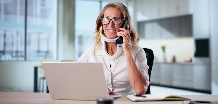 A woman wearing a neck brace smiles while talking on the phone with the Law Offices of Humberto Izquierdo Jr. PC at her desk in front of a laptop suggesting she is following up on a workers' comp claim while continuing to manage her daily responsibilities.