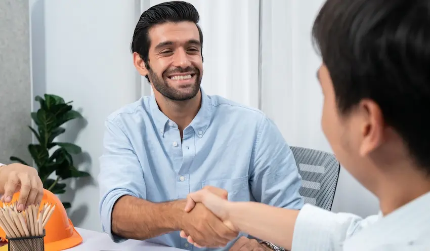 A smiling man in a light blue shirt shakes hands across a desk with an orange hard hat nearby representing the successful resolution of a workers' comp claim with the help of a knowledgeable attorney from Law Offices of Humberto Izquierdo Jr. PC in Georgia.
