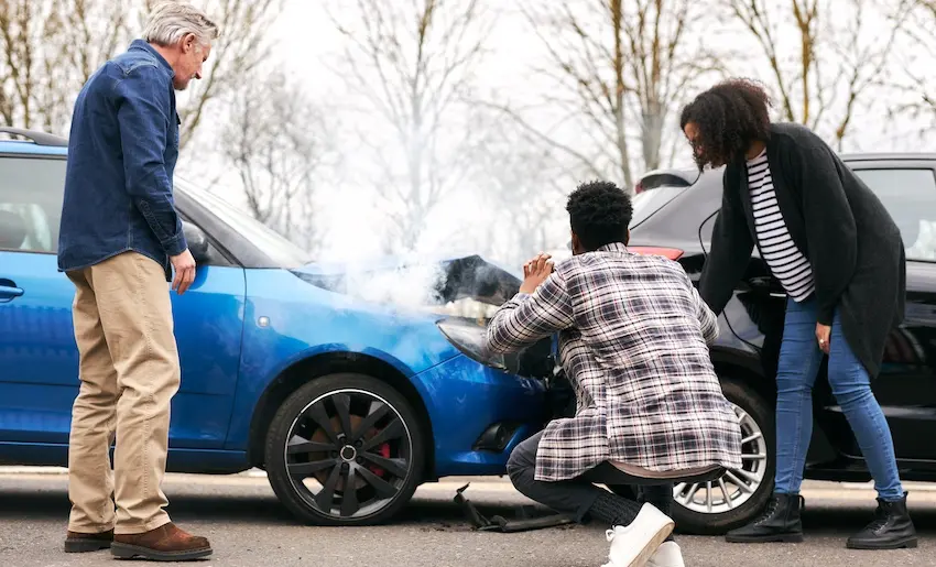 Three people gather around the crumpled front end of a blue sedan on a gray overcast day with white smoke rising from the damaged hood of the vehicle — an older man with white hair in a denim jacket and khaki pants stands upright on the left looking down at the damage- a young man in a plaid flannel shirt crouches down in the center pressing his hands together as he examines the crushed bumper up close and a Black woman with curly hair wearing a black cardigan over a striped shirt stands on the right leaning in to assess the damage — depicting the tense moments at an accident scene before victims begin the process of contacting their insurance company to file a claim in Georgia.