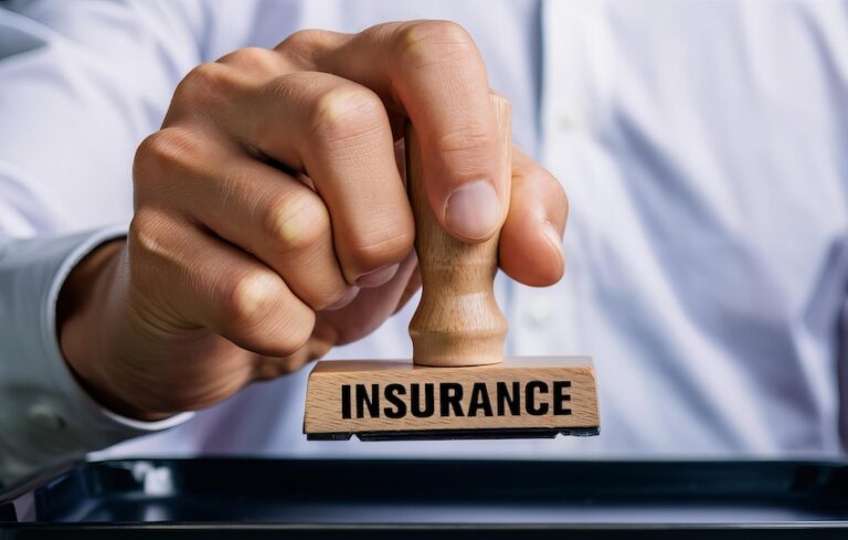 A close-up of a person's hand gripping a wooden rubber stamp with a flat rectangular base bearing the word INSURANCE printed in bold black capital letters pressing it firmly down onto a dark ink pad on a reflective black surface with the person wearing a crisp white dress shirt with the cuff visible at the wrist — representing the formal approval and documentation process carried out by an insurance company when processing a policyholder's claim" in Georgia.
