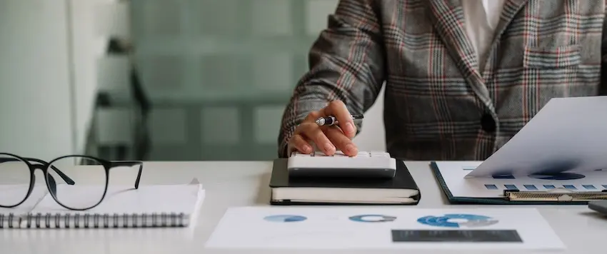 An attorney from the Law Offices of Humberto Izquierdo Jr. PC is wearing a gray and red plaid blazer over a white top leans forward at a clean white desk using one hand to operate a small white desktop calculator while holding a black and silver pen in the other with a spiral-bound white notepad- a pair of black-framed reading glasses- a dark hardcover notebook- and two printed documents featuring blue pie charts and circular data graphics spread across the desk in front of them against a blurred glass partition office background — illustrating the detailed financial analysis and policy review work performed by an insurance company adjuster or claims specialist