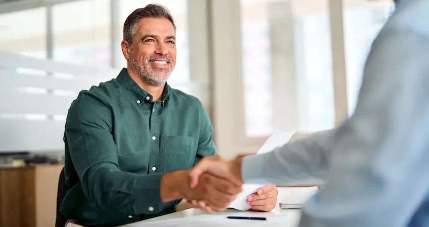 A middle-aged man with short salt-and-pepper hair and a neatly trimmed beard wearing a dark teal button-down shirt sits at a white table smiling broadly as he reaches across to shake hands with a lawyer from the Law Offices of Humberto Izquierdo Jr. PC visible only from behind in a light blue shirt who is simultaneously handing over a document with a hardcover book and a blue pen resting on the table between them and large frosted windows flooding the modern office space with natural light — capturing the moment of resolution and agreement when a client successfully settles a claim with their insurance company in Georgia.